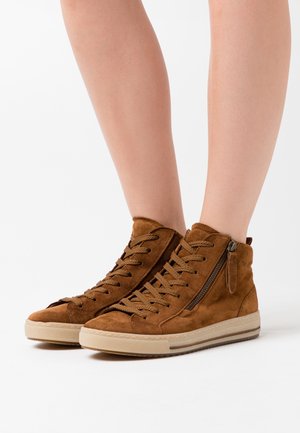 Bruine suède hoge sneakers met veters en zijritsen, gedragen op blote benen tegen een witte achtergrond.