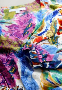 Buntes Blumenmusterstoff mit lila, blau, grün, orange und weißen Mustern, zeigt eine Nahaufnahme eines gerafften Ärmels und eines Teils eines Halsausschnitts.