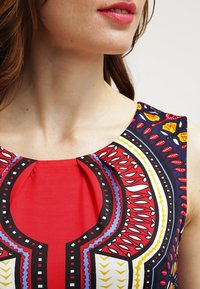 Ženský výstřih a rameno, oblečené v barevném vzorovaném topu bez rukávů s geometrickými motivy v červené, černé, modré, žluté a bílé barvě.