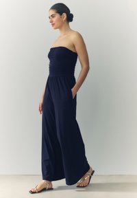Marine jumpsuit med en rynket, uden skulderstropper bodice, brede bukser og sidelommer. Parret med guld flade sandaler. Glat, letvægtsstof.