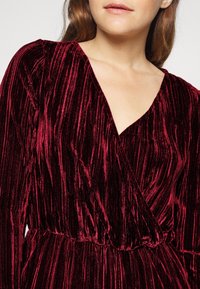 Šaty z vínově červeného sametu s vertikálním žebrovaným vzorem, výstřihem do V, dlouhými rukávy a detailem s nařaseným pasem. Luxusní a jemný vzhled.