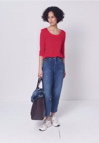 Femme aux cheveux bouclés portant un haut rouge, un jean bleu, des baskets blanches, tenant un sac fourre-tout sombre avec une veste en denim pliée à l'intérieur.