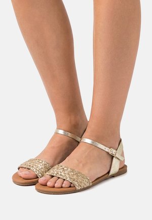 Pies con sandalias planas metálicas doradas con tiras tejidas en los dedos y correas con hebilla en el tobillo, sobre un fondo gris claro.