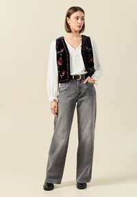 Gilet di velluto nero con ricami floreali sopra una camicia bianca, abbinato a jeans grigi a vita alta e pantaloni larghi e zoccoli neri.