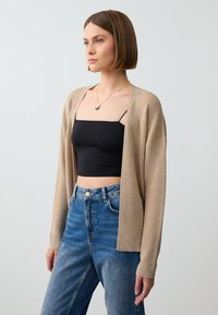 Beige Strickcardigan mit langen Ärmeln, offenem Frontbereich und gerippten Bündchen, getragen über einem schwarzen figurbetonten Oberteil und blauen hochtaillierten Jeans.