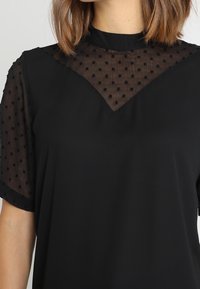 Top negro con mangas cortas y transparentes con puntos, y un escote texturizado y semitransparente. Presenta un corte holgado y un diseño en forma de V sutil en el pecho.