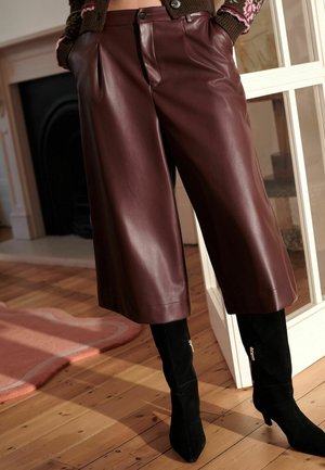 Burgunderfarbene, aus Kunstleder gefertigte Culottes mit Falten an der Taille, kombiniert mit knielangen schwarzen Wildlederstiefeln mit goldenen Reißverschlüssen.
