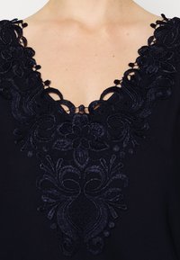 Blusa blu navy con ricami floreali intricati sul collo, caratterizzata da una ricca texture e delicati dettagli a intaglio.
