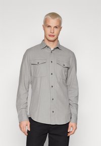 Grå långärmad skjorta i texturerat tyg, med två bröstfickor, knäppning framtill och rundad nederkant med subtila ränder.