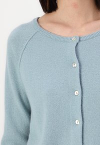 Hellblauer Cardigan mit einer weichen, gestrickten Textur, rundem Halsausschnitt und einer Vorderknopfleiste mit vier weißen Knöpfen.