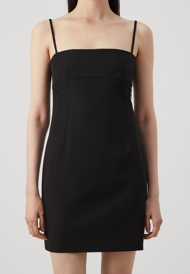 Femme portant une mini robe noire sans manches avec des fines bretelles sur un fond clair uni.