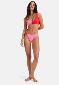 Two-tone bikini set in textured fabric; top features a twist design with red and pink sections, matching pink bikini bottom with minimal coverage.
