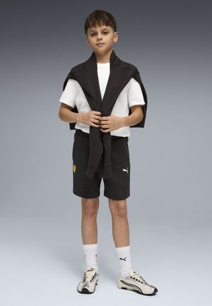 Dreng stående med sort sweatshirt bundet om skuldrene, iført hvid T-shirt, sorte Puma-shorts, hvide Puma-sokker og hvide sneakers.