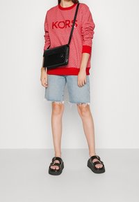 Felpa rossa con motivo a fantasia e lettering KORS, pantaloni shorts di jeans, borsa a tracolla nera e sandali neri chunky con fibbie e suole testurizzate.