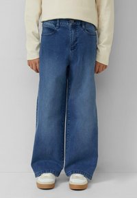 Persoon in losse blauwe denim jeans, witte sneakers met rubberen zolen en een lichtbeige langemouwen top, staande tegen een effen muur.