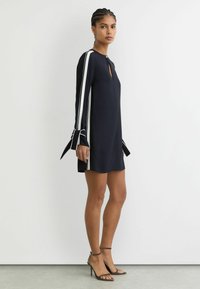 Tumši zila kleita ar baltām sānu svītrām, garām piedurknēm ar loku akcentiem un izgriezumu pie kakla. Gluds audums un ērtas valkāšanas piegriezums.