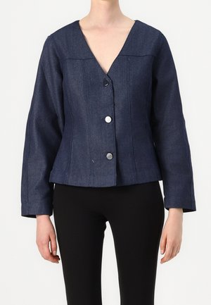 Kobieta w ciemnoniebieskiej, dżinsowej kurtce z długim rękawem, z trzema dużymi srebrnymi guzikami, w czarnych spodniach na jednolitym tle.