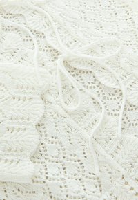 Haut en tricot en dentelle blanc, présentant un motif texturé avec des bords festonnés et une fermeture à lien central en fil assorti.