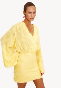 Vestido amarillo con un tejido texturizado y deshilachado. Presenta un escote en V profundo, mangas largas y una falda ajustada. Combinado con pendientes en forma de cereza.