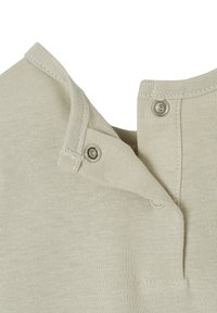Helles beiges Baumwollkleidungsstück mit einem Druckknopfverschluss an der Schulter, das eine weiche Textur und saubere Nähdetails aufweist.