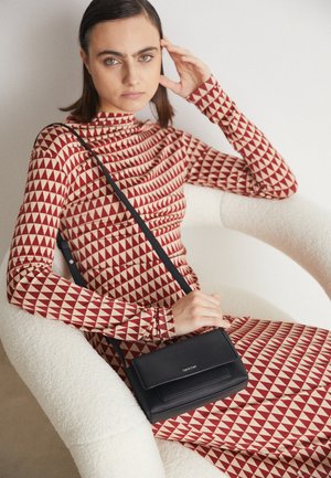 Vrouw met steil donker haar die een rood-beige jurk met geometrisch patroon draagt, zittend in een witte, getextureerde stoel, houdend een zwarte crossbodytas.