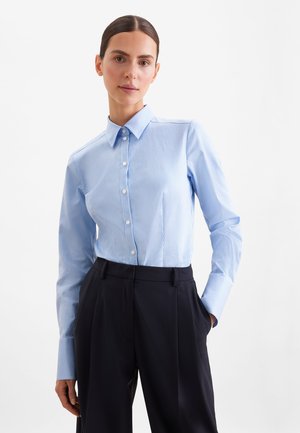 Kvinde med mørkt hår samlet bagpå, iført en lyseblå stribet skjorte med knapper og sorte bukser med høj talje, stående med den ene hånd i lommen.