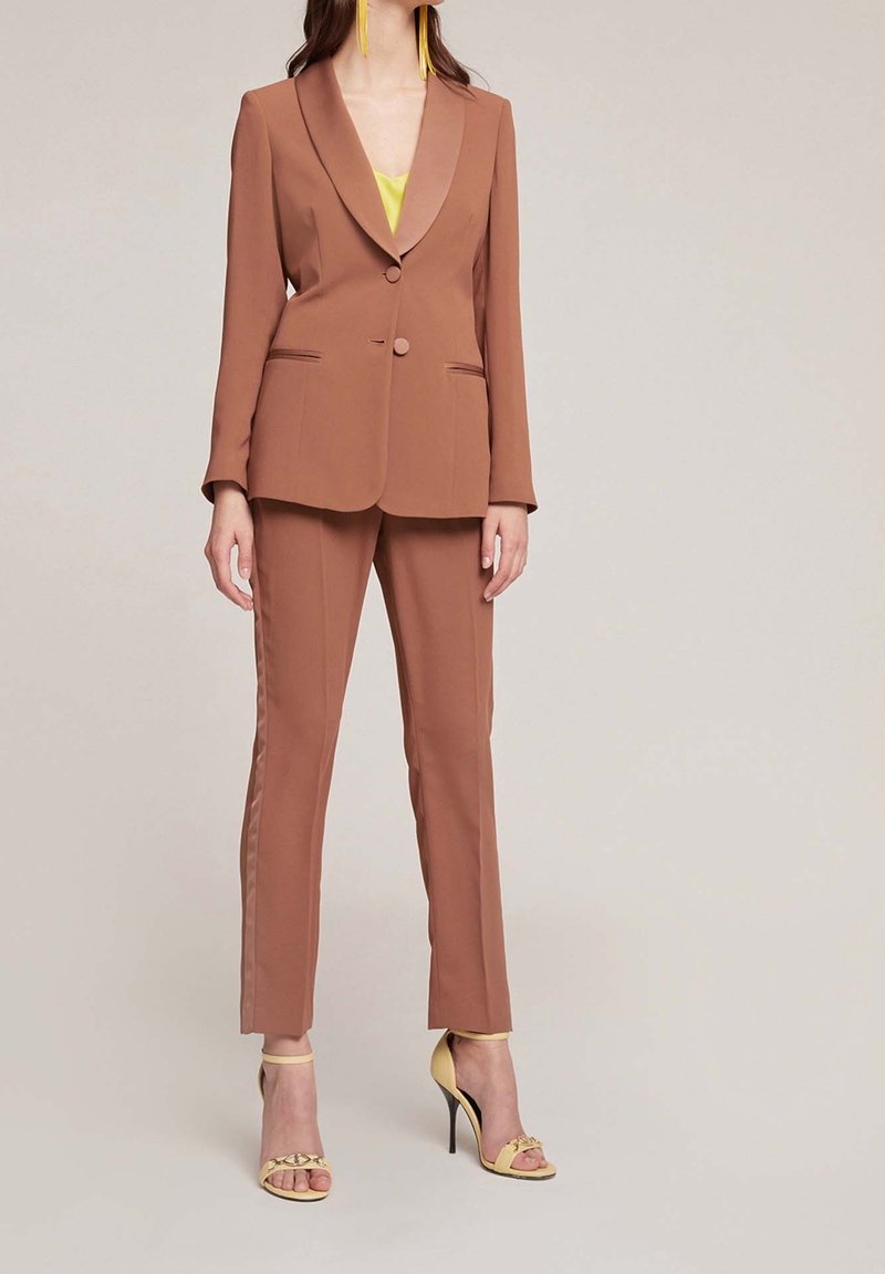 Hnědý oblek na míru s klopami, jednořadým zapínáním na knoflík, bočními kapsami a kontrastními pruhy na kalhotách. Nosený s žlutými podpatky.
