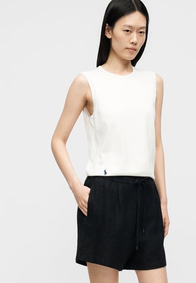 Γυναίκα με μακριά μαύρα μαλλιά φοράει λευκό αμάνικο τοπ και μαύρα σορτς με κορδόνι, στέκεται με το ένα χέρι στην τσέπη σε απλό φόντο.