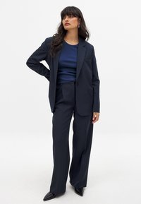 Námornícky modrý oblek na mieru so sakom na jednoročný gombík a širokými nohavicami, doplnený tmavomodrým rebrovaným tričkom a čiernymi špicatými lodičkami.