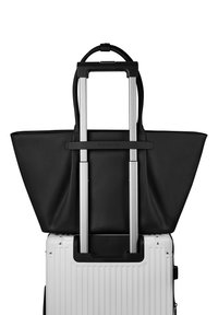 Grand sac cabas en cuir noir accroché à la poignée télescopique d'une valise rigide blanche à roulettes sur fond blanc.