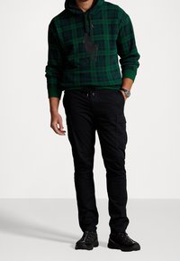 Zelená kostkovaná mikina s kapucí s černými doplňky a logem, doplněná černými cargo kalhotami. Texturovaná látka a volný střih. Černé tenisky.