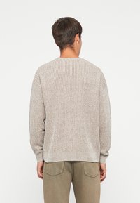 Lichtbeige gebreide trui met een geribbelde textuur, lange mouwen en een ronde hals, van achteren weergegeven. Casual pasvorm, minimale details.