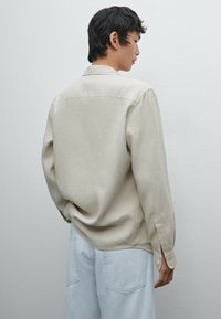 Beige long-sleeve shirt with a collar, slightly relaxed fit, and buttoned cuffs, paired with light blue denim pants. Minimalist design.
