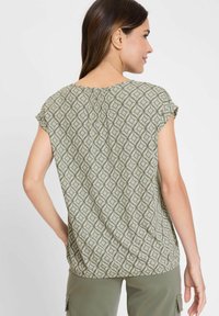 Grün gemusterte Bluse mit kurzen Ärmeln, die ein wellenartiges Design und Raffungen am hinteren Halsauschnitt aufweist. Aus leichtem Stoff gefertigt.