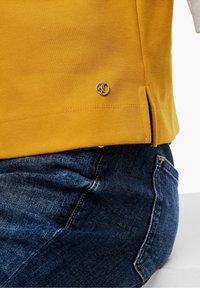 T-shirt en coton jaune moutarde avec une fente latérale et un logo doré au niveau de l'ourlet, associé à un jean en denim foncé montrant un tissu texturé.