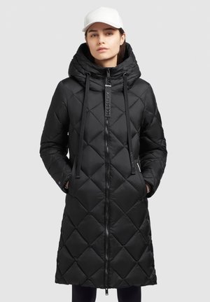Femme portant un long manteau noir matelassé avec capuche et cordons de serrage, fermeture éclair à l'avant, les mains dans les poches, et une casquette de baseball blanche.