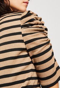 Top de manga abullonada en beige con rayas horizontales negras. Presenta una tela texturizada con un sutil efecto arrugado, exhibiendo un diseño de hombro fruncido.