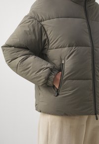 Person in olivgrünem Daunenjacke mit Hand in der seitlichen Reißverschlusstasche, kombiniert mit cremefarbenen Cordhosen vor einem neutralen Hintergrund.