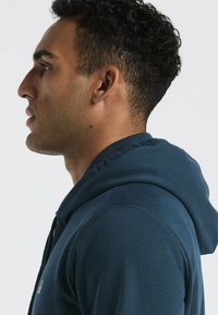 Blauwe capuchontrui met een geribbelde textuur, naaddetails en een klein logoaccent op de borst. Zijkant profielweergave.