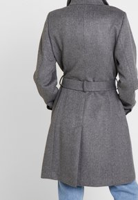 Cappotto grigio in lana con vita cintata, maniche lunghe e spacco posteriore. La texture è liscia con un design di sartoria classico.
