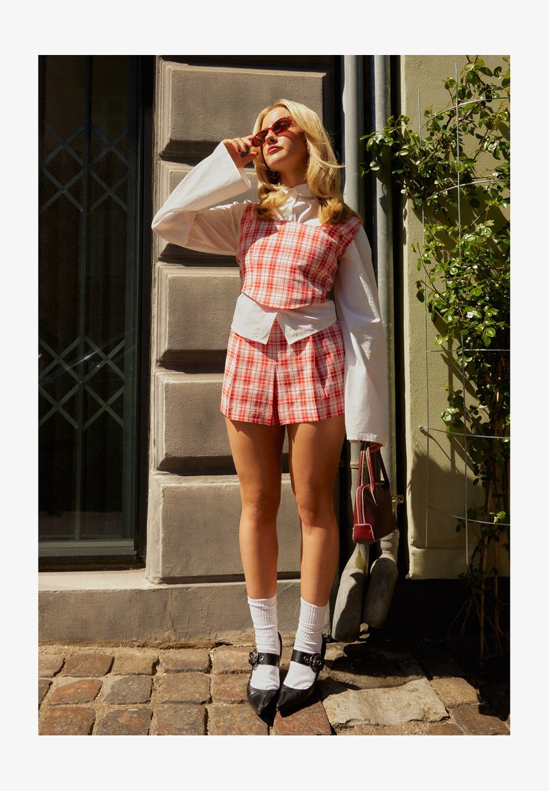 Ensemble de top court à carreaux rouges et blancs avec un haut blanc en dessous. Des ballerines noires à bouts pointus avec détail de boucle. Les accessoires incluent des lunettes de soleil rouges et un sac à main.