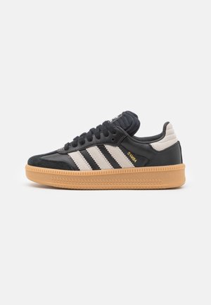 Schwarzer und beiger Adidas Samba Sneaker mit Gummisohle und drei beigen Streifen, seitlich auf weißem Hintergrund betrachtet.