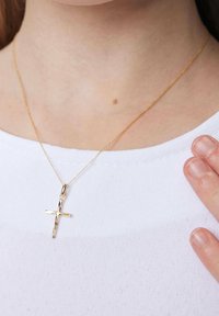 Złoty wisiorek krzyż na cienkim łańcuszku, z fakturowaną powierzchnią. Noszony na gładkiej białej bluzce, z ręką delikatnie dotykającą naszyjnika.