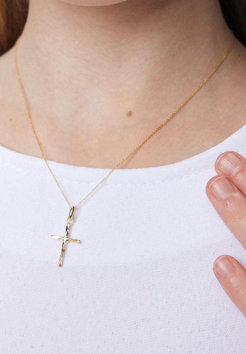 Złoty wisiorek krzyż na cienkim łańcuszku, z fakturowaną powierzchnią. Noszony na gładkiej białej bluzce, z ręką delikatnie dotykającą naszyjnika.