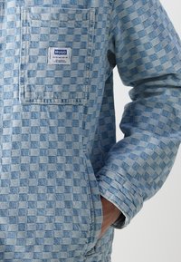 Woven check-patterned jacket in light blue denim with a front pocket, textured finish, and visible stitching details.