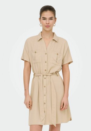 Donna che indossa un vestito beige a maniche corte con bottoni, tasche sul petto e cintura annodata in vita, in piedi davanti a uno sfondo neutro.