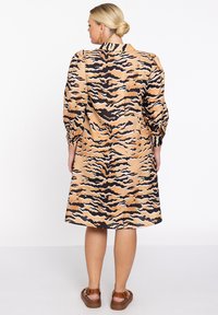 Lange mouw jurk in tan stof met een zwart en bruin tijgerstreep patroon, losse pasvorm en een halflange zoom. Bruine sandalen maken de look compleet.