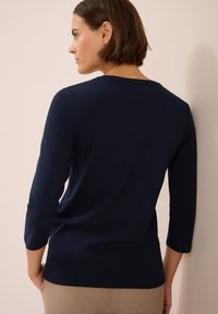 Frau mit kurzen braunen Haaren, die ein langärmliges navyblaues Shirt trägt, nach links blickend, vor einem neutralen beige Hintergrund.