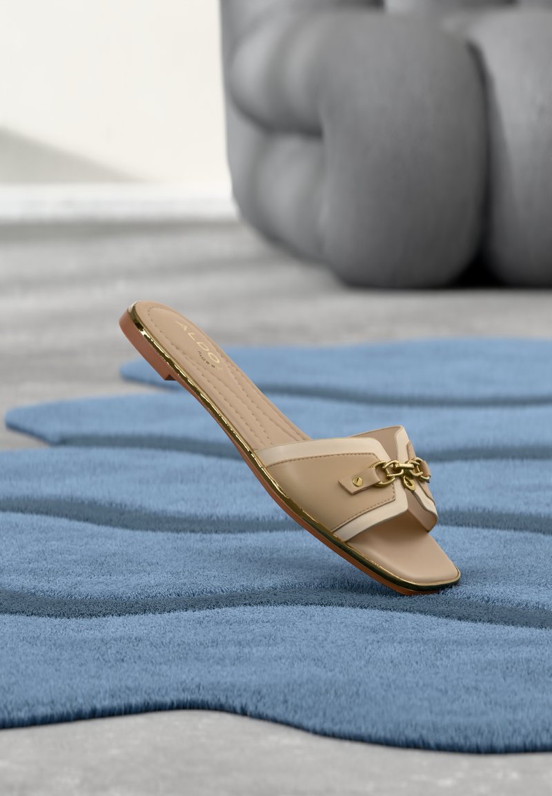Sandale plate beige à bout ouvert avec un détail en chaîne dorée reposant sur un tapis bleu texturé, avec un coussin gris flou en arrière-plan.
