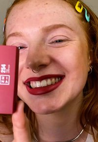 Röd läppstift i en rektangulär låda märkt "254" som hålls upp av en leende person; utmärker sig med livfull färg, slät textur och minimalistisk design.